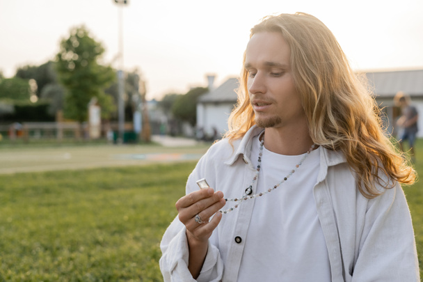 thoughtful yoga man in white linen shirt looking at crystal on mala beads while standing in countryside outdoors - Foto, afbeelding