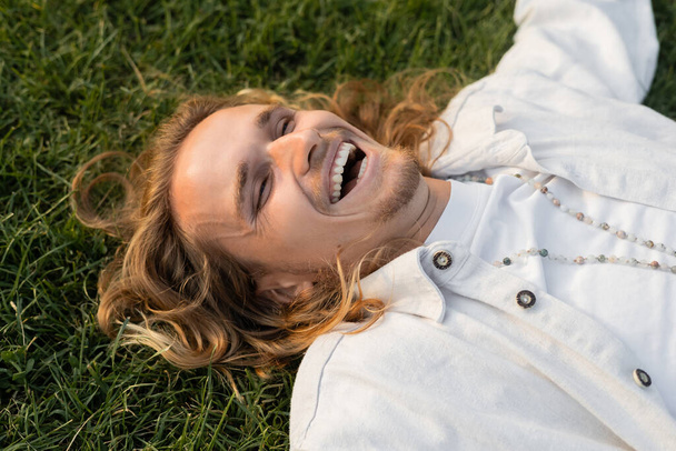 άποψη υψηλής γωνίας του ευτυχισμένος άνθρωπος σε λευκό πουκάμισο και χάντρες που βρίσκονται σε πράσινο γρασίδι σε εξωτερικούς χώρους - Φωτογραφία, εικόνα