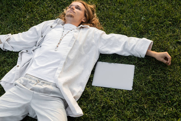magas szög kilátás fiatal jóga férfi fehér ruhák közelében fekvő laptop zöld mezőn  - Fotó, kép