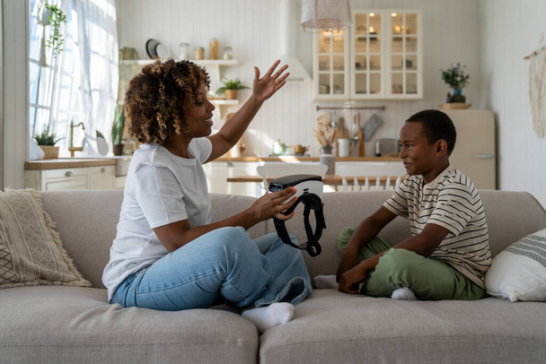 Young African American woman mother sitting on sofa with little boy son holding vr headset talking about metaverse opportunities with kid. Immersive virtual reality and developing child - Foto, afbeelding