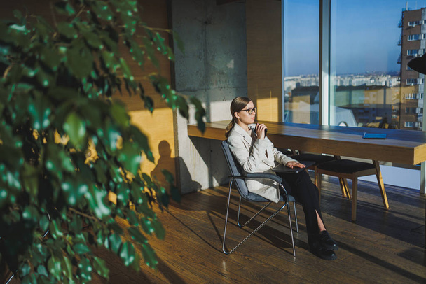 Улыбающаяся молодая деловая женщина в повседневной одежде и очках сидит в офисе с мобильным телефоном во время работы над удаленным проектом с помощью ноутбука - Фото, изображение