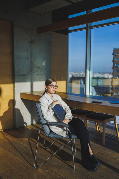 Lachende jonge zakenvrouw in casual kleding en bril zittend in het kantoor met een mobiele telefoon tijdens het werken aan een project op afstand met behulp van een laptop - Foto, afbeelding