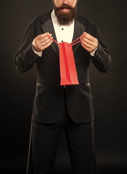 surprised curious man in tuxedo bow tie formalwear on black background with business reward in shopping bag. occasion greeting. - Foto, Imagen