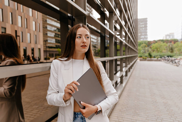 Buitenfoto van stijlvolle Europese vrouw met los haar in witte jas en jeans houdt laptop vast en kijkt opzij terwijl ze poseert in de stad op de achtergrond van een modern gebouw - Foto, afbeelding