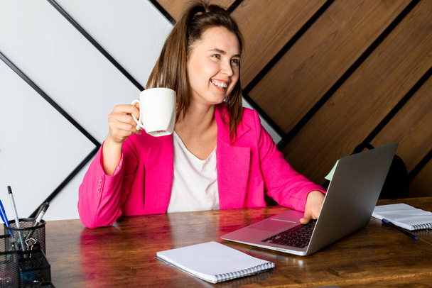 Portret uśmiechniętej pracowniczki biurowej lub pracownika IT siedzącego przy biurku w nowoczesnym biurze z laptopem i filiżanką w dłoni. - Zdjęcie, obraz