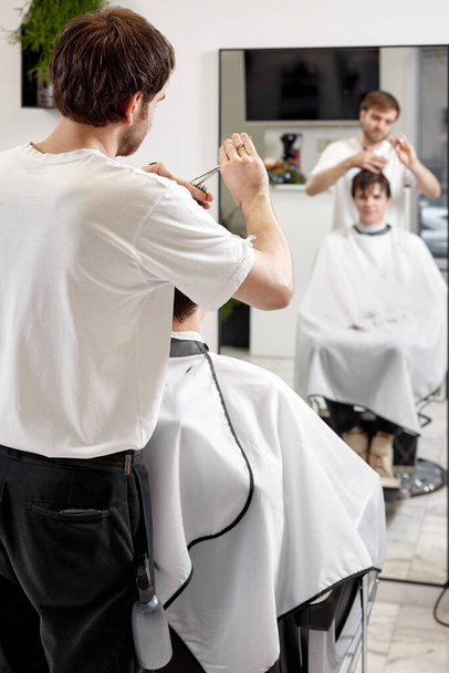 young caucasian man getting haircut by professional male hairstylist using comb and grooming scissors at barber shop. - Фото, зображення