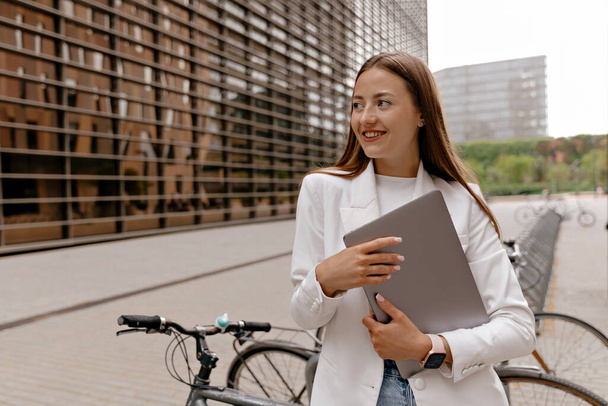 Beyaz ceketli, kot pantolonlu, mutlu gülüşlü sevimli bir kız elinde dizüstü bilgisayarla modern binaların ve şehir bisikletlerinin arka planında kameraya poz veriyor. - Fotoğraf, Görsel