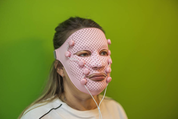 Close-up uitzicht op de vrouw met roze gezicht massager masker op groene achtergrond. Zweden.  - Foto, afbeelding