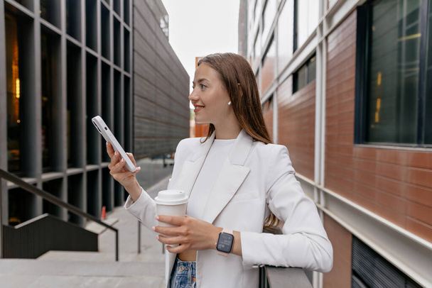 Profile outdoor portrait of pretty stylish girl in white jacket and top is using smartphone and drinking coffee in city. Young lady staying near to wall with phone. Street style concept. - Photo, Image