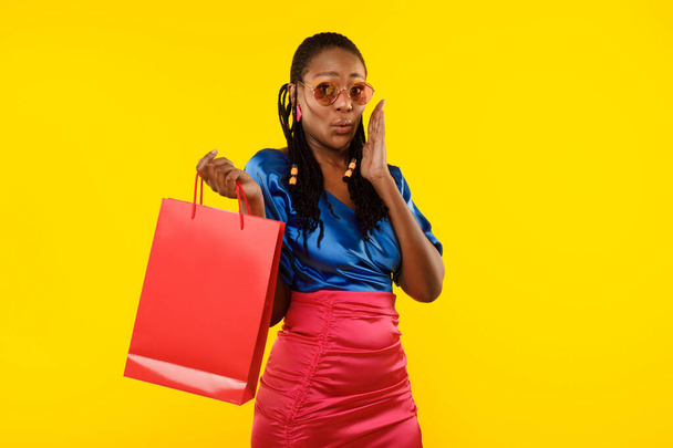 Шопінг. Стильна чорна жінка позує в сонцезахисних окулярах, одягнених в елегантний одяг і тримає червону паперову сумку, що стоїть на жовтому фоні студії. Пропозиція літніх продажів
 - Фото, зображення