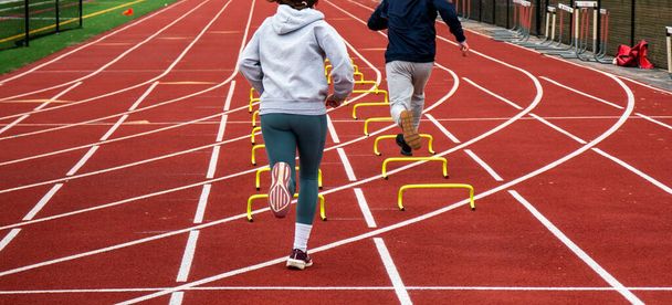 Visão traseira de dois corredores correndo mais de seis polegadas mini barreiras amarelas em pistas em uma pista no treino. - Foto, Imagem