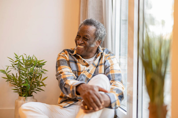 Счастливый пенсионер сидит на полу дома рядом с окном - Фото, изображение