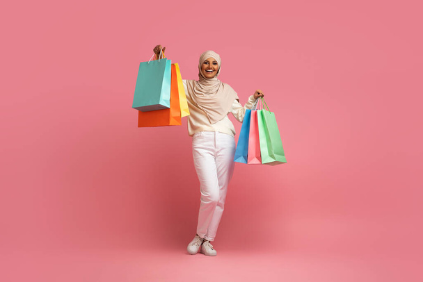 Concepto de ventas estacionales. Mujer musulmana emocionada en hijab sosteniendo un montón de bolsas de compras, dama shopahilic islámica llevando compras y sonriendo a la cámara, de pie sobre fondo de estudio rosa - Foto, Imagen