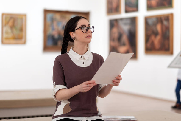 Portré kaukázusi csinos fiatal nőről, aki szemüveget visel, és oktatási brosúrát tanul. Defokted képek a háttérben. A kiállítás koncepciója a galériában. - Fotó, kép