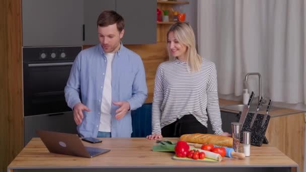 Agréable couple familial debout près de grande table en bois dans la cuisine moderne, regardant l'écran d'ordinateur portable. Heureux jeune couple commence à préparer le déjeuner. - Séquence, vidéo