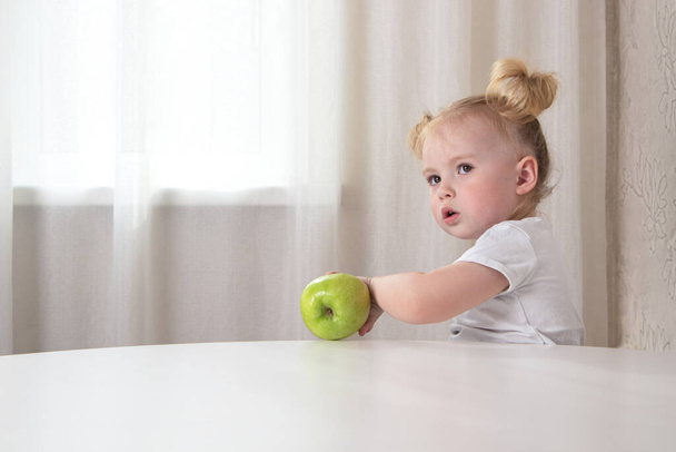 白いTシャツにポニーテールの女の子は、彼女の手に緑のリンゴとテーブルに座っています - 写真・画像