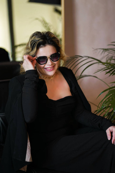 A smiling woman in a black suit and dark glasses sits on a rug. - Foto, afbeelding