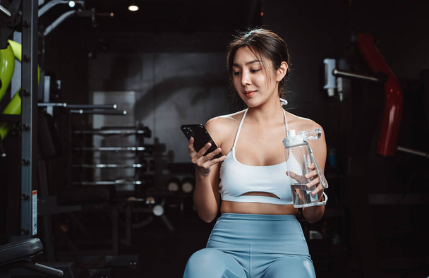 Woman checking the time and using smartphone exercise workout in gym fitness breaking relax after training sport with dumbbells and water bottle healthy lifestyle bodybuilding. - Foto, Imagen