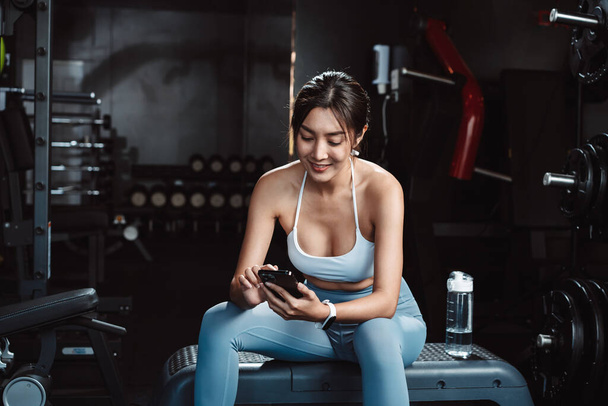 Woman checking the time and using smartphone exercise workout in gym fitness breaking relax after training sport with dumbbells and water bottle healthy lifestyle bodybuilding. - Φωτογραφία, εικόνα