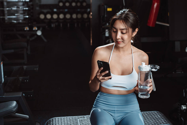 Femme vérifiant le temps et utilisant l'exercice smartphone séance d'entraînement dans la salle de gym fitness rupture se détendre après l'entraînement du sport avec haltères et bouteille d'eau mode de vie sain musculation. - Photo, image