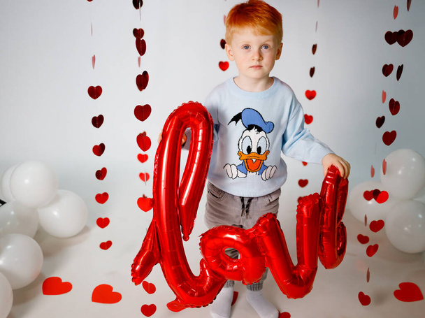 Un ragazzo dai capelli rossi su uno sfondo bianco con le palle rosse. Abbigliamento elegante per bambini - Foto, immagini