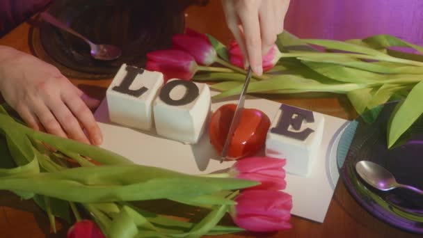 Hintergrund der Torten mit dem Wort Liebe zwischen frischen Tulpen auf dem Serviertisch, Hände, die das Herz in zwei Stücke schneiden. Liebesbotschaft, Valentinstag, unerwartete Überraschung, HQ 4k Filmmaterial - Filmmaterial, Video