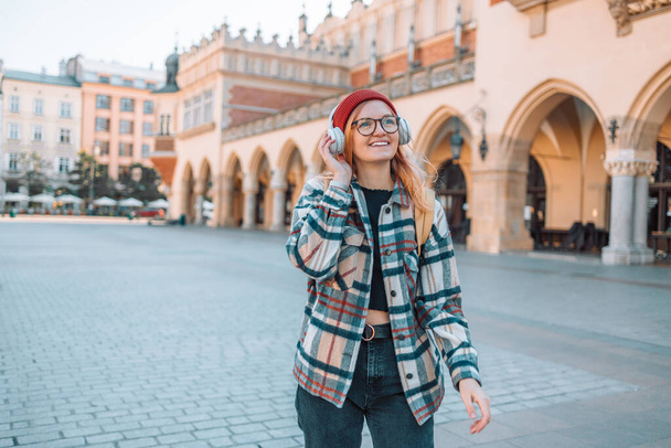 Jonge blonde vrouw glimlachen blij met behulp van smartphone en koptelefoon, poseren in de straat van de Europese Krakau stad. Hoge kwaliteit foto - Foto, afbeelding