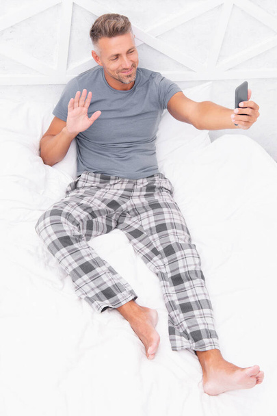 video call man with phone in bed gesturing hello. photo of video call man with phone at home. video call man with phone in bedroom. video call man with phone wear pajama. - 写真・画像