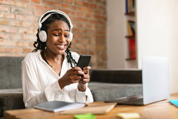Портрет усміхненої чорної жінки, що сидить за столом з ноутбуком, використовує мобільний телефон і носить навушники, вільний простір. Щаслива леді в бездротовій гарнітурі дивитися відео або спілкуватися перегляд соціальних медіа
 - Фото, зображення