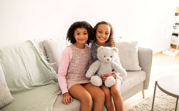 Две симпатичные афроамериканские сестры обнимаются с пушистым медвежонком, сидя на диване, позируя вместе в повседневных платьях дома, улыбаясь в камеру. Концепция дружбы братьев и сестер - Фото, изображение