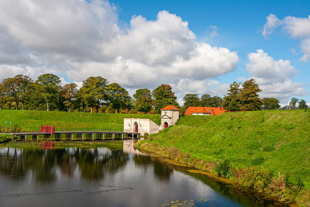 Copenhagen, Denmark - September 13, 2010: Wide green landscape under blue cloudscape with green ramparts, bridge and south gate into Kastellet, citadel. Red roofs peep over, dark water - Photo, Image