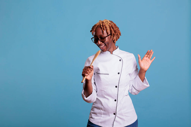 Amerykański kucharz afrykański portret śpiewający z naczyniem kuchennym. Kobieta profesjonalna kuchenka dobrze się bawi, śmieje, śpiewa piosenkę. uśmiechnięta kuchnia pracownik średni strzał korzystających. - Zdjęcie, obraz