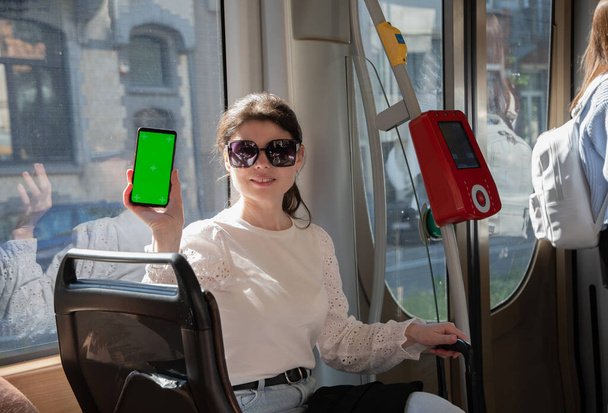 morena mujer mostrando smartphone con pantalla verde vertical en tranvía, junto a la caja digital Foto de alta calidad - Foto, Imagen