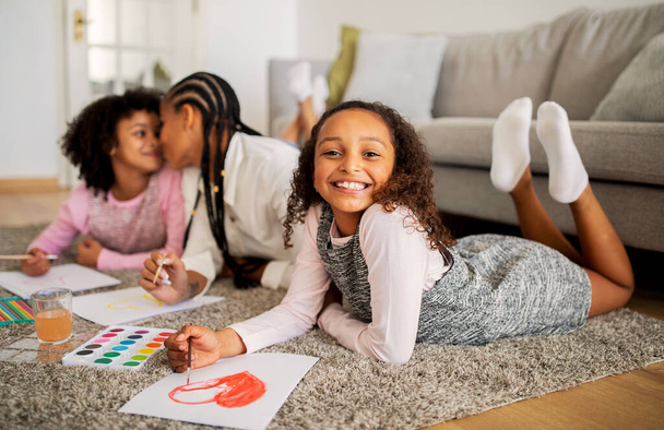 Ευτυχισμένο μαύρο κοριτσάκι ζωγραφίζει με τη μητέρα και την αδελφή μαζί, δένεται και διασκεδάζει ξαπλωμένο στο πάτωμα του σαλονιού στο σπίτι, χαμογελώντας στην κάμερα. Οικογενειακό Σαββατοκύριακο Leisure Concept. Βάθος κλίσης - Φωτογραφία, εικόνα