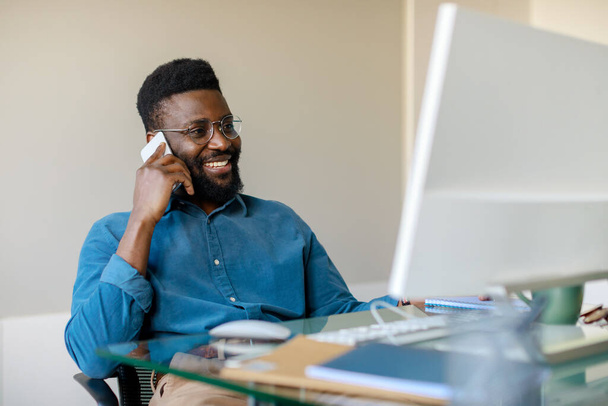 Gelukkige zwarte zakenman die een telefoongesprek heeft met een cliënt, achter de computer op kantoor zit en praat op een mobiele telefoon. Modern bedrijfsconcept - Foto, afbeelding