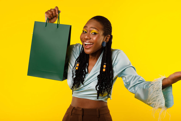 Bevásárlási ajánlat. Vidám fekete nő mutatja zöld könyv Shopper táska pózol fényes smink mosolygott a kamera felett álló sárga háttér stúdióban. Portré vidám Shopaholic Woman - Fotó, kép