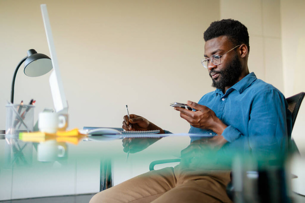 Σχέδιο διαχείρισης χρόνου. Σοβαρός μαύρος επιχειρηματίας που χρησιμοποιεί κινητό τηλέφωνο και κρατάει σημειώσεις, διαχειρίζεται σχέδια ενώ εργάζεται στο γραφείο, ελεύθερος χώρος - Φωτογραφία, εικόνα