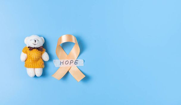 One yellow ribbon with sticky tape with hope and a toy bear lie on the left on a blue background with copy space on the right,flat lay close-up.Concept for World Childhood Cancer Day. - Fotografie, Obrázek