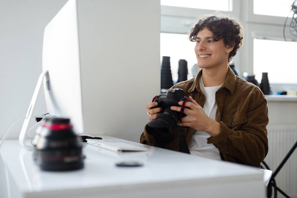 Szczęśliwy profesjonalny fotograf trzymający kamerę i patrzący na monitor komputerowy, siedzący w miejscu pracy w studio. Profesjonalny artysta przesyłający zdjęcia do komputera w celu retuszu - Zdjęcie, obraz