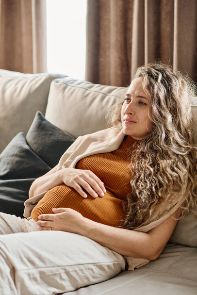 Mujer embarazada con el pelo largo y ondulado rubio manteniendo las manos en su vientre mientras se relaja en un sofá suave y viendo la televisión en el ocio - Foto, Imagen