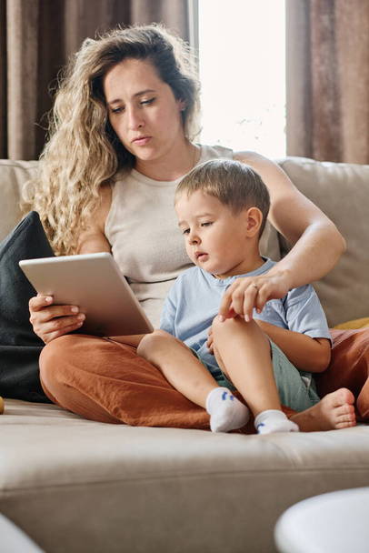 Young serious blond woman with tablet and her cute little son watching online movie for children or cartoons while sitting on sofa - Φωτογραφία, εικόνα