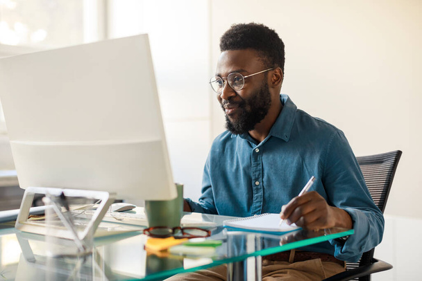 Mutlu Afro-Amerikan girişimci, çalışır vaziyette bilgisayar ekranına bakıyor, ofiste notlar alıyor, yeni iş fırsatları arıyor, yer kopyalıyor. - Fotoğraf, Görsel