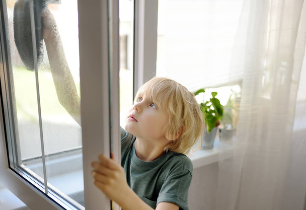 Cute little boy washing a window at home. Child helping parents with household chores, for example, cleaning windows in his house. Children doing housework. Kids household duties - Zdjęcie, obraz