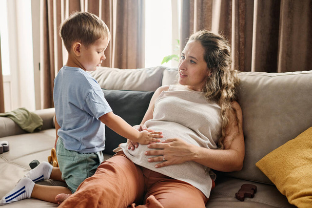 Curious toddler keeping hand on belly of his pregnant mother sitting on sofa while spending spare time with her cute little son at home - Foto, imagen