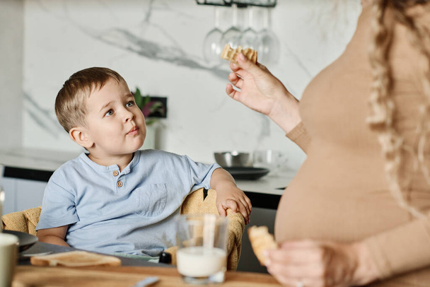 Cute little boy looking at his pregnant mother with piece of homemade cookie in hand while having breakfast by kitchen table - Foto, Imagen