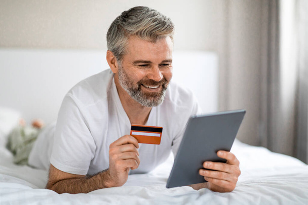 Homme mûr aux cheveux gris attrayant et souriant portant un pyjama profitant des achats en ligne tout en se relaxant le week-end à la maison, allongé sur le lit, tenant une tablette numérique et une carte bancaire, espace de copie - Photo, image