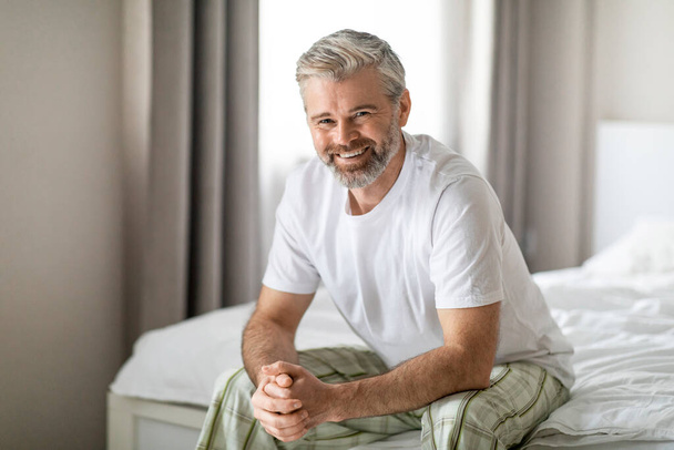 Retrato de feliz atraente de cabelos grisalhos de meia idade homem vestindo pijama sentado na cama em quarto branco confortável em casa, sorrindo para a câmera, desfrutando de bom dia após o sono adequado - Foto, Imagem
