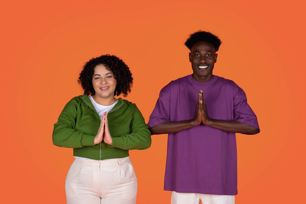 Relaxation, stress relief, meditation. Cheerful beautiful young multiracial couple folded their hands in front of them and smiling, showing namaste gesture, red studio background - Photo, Image