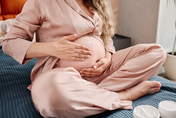 Mulher grávida em pijama de seda aplicando creme hidratante em sua barriga antes de dormir ou após o banho enquanto sentada em cama de casal - Foto, Imagem