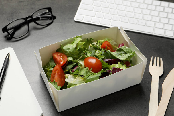 Container of tasty food, keyboard, glasses, cutlery and notebook on grey table. Business lunch - Photo, image